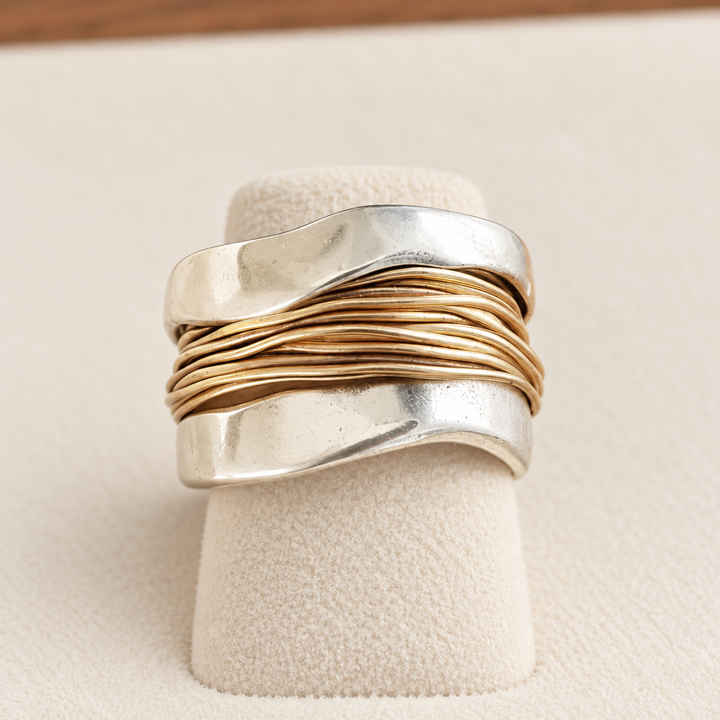 Anillo ancho plateado con hilos dorados entrelazados, sobre un soporte de terciopelo crema
